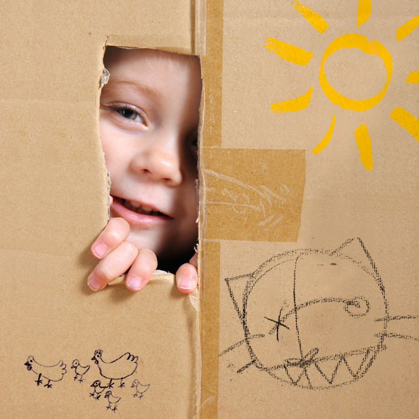 Kind spitzt durch Fenster eines Kartonhauses.