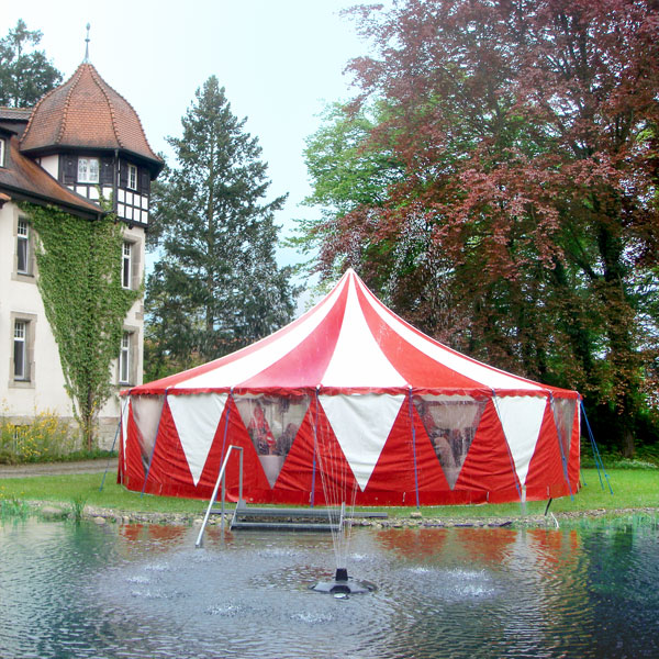 Rot-weißes Zirkuszelt steht vor einem Schloss und einem See.