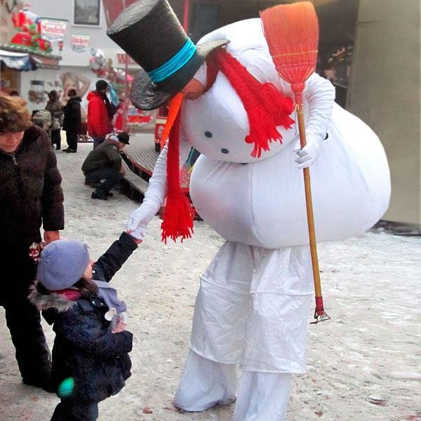 Schneemann-Stelzenläufer begrüßt auf dem Weihnachtsmarkt ein Kind..