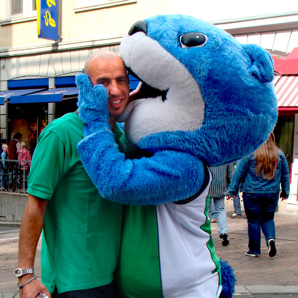 Blaues Maskottchen hält Promoter im grünen Shirt im Arm.
